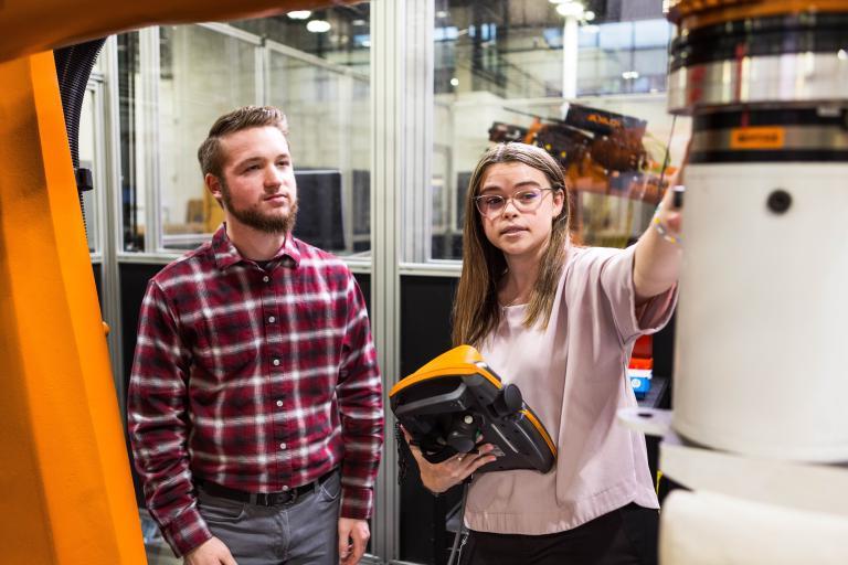 Two Kettering University students, a female and a male, look at a large piece of machinery.